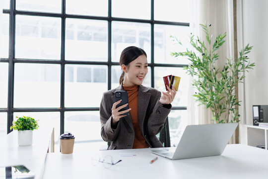 Asian woman making online payment with two credit cards and mobile phone while sitting at office desk, using laptop for e-commerce shopping and payment processing