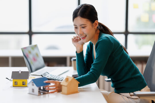 Young asian woman selecting a house miniature model, contemplating real estate investment options, making a decision on choosing a new home, and planning future property ownership