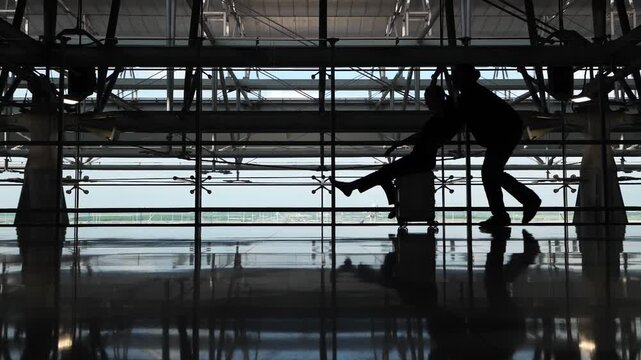 Cheerful couple of tourists have nothing to do but ride their suitcases around the empty terminal. Two passengers pass the time before boarding a flight, having as much fun as they can