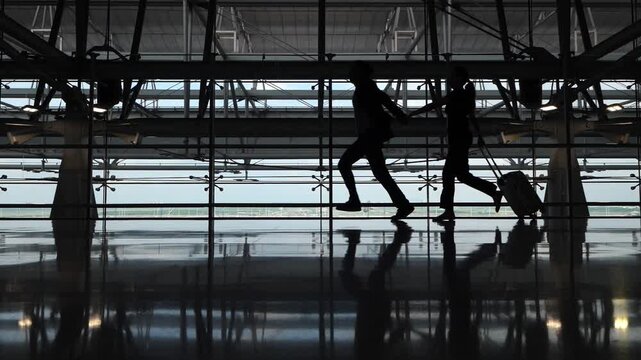Misguided couple of tourists run through terminal to boarding gate, slow motion silhouetted shot. They are afraid of missing their flight and are in hurry, man pulls girl by hand