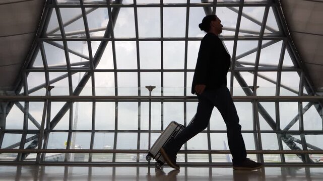 Passenger go to boarding gate, side view, slow motion. Man rolling a small suitcase behind him, dark figure against glass wall of modern airport. Guy with hipster hair wear casual suit