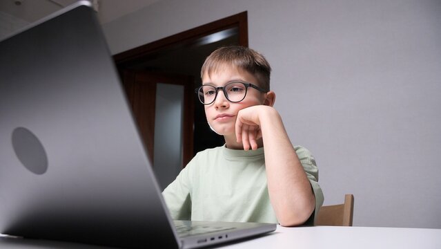 Young boy with glasses feeling tired and bored, resting his chin on his hand while looking at a laptop screen during remote learning or online education at home