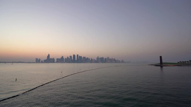 Panoramic dusk shot of Doha's West Bay skyline seen from MIA Park, with calm reflective waters, a curved breakwater, and a soft orange and purple gradient sky silhouetting the modern towers.
