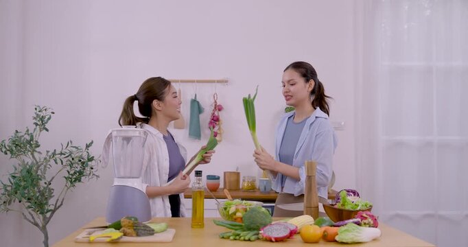 Asian LGBTQ female couple stands at kitchen counter, holds fresh scallion and cucumber, chats and smiles during holiday meal prep, cozy home cooking routine, playful bonding, healthy weekend love