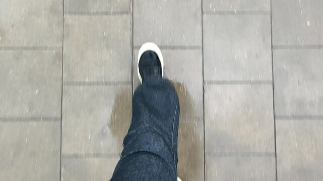 POV top down view of feet in jeans and sneakers walking on wet pavement in the rain. The reflection of an umbrella is visible on the shiny wet tiles below. Concept of point of view, walking