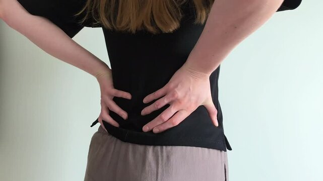Woman grips her lower back with both hands suffering from sharp pain. She holds her spine tightly while bending slightly forward in discomfort. Concept of severe backache, muscle spasm and