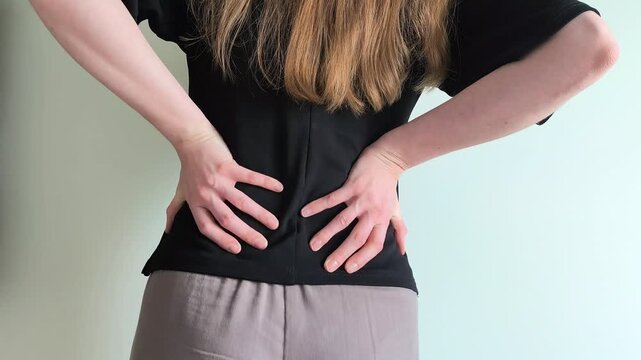 Woman places both hands on her lower back showing discomfort and pain. She holds her spine while standing reacting to lumbar tension. Concept of backache, posture problems and lumbar muscle