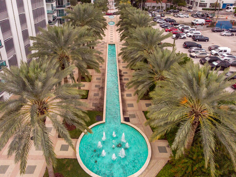 Miami, Florida, United States - 24 December 2019: Aerial view of a long turquoise fountain lined with rows of palm trees in a park, with a nearby parking lot and urban buildings.