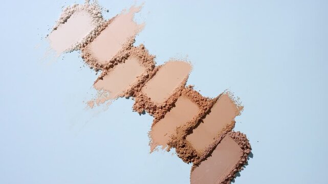 Vertical crane view of seven from beige to brown loose powder streaks arranged linearly on a pale blue surface rotating directional light produces ever-changing shadows