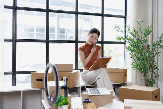 Female entrepreneur managing small parcel delivery, talking on smartphone, checking order details on clipboard in her home office, building a successful online business
