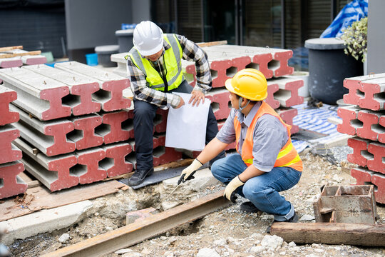 A skilled builder and engineer work on the precise alignment of a foundation beam. A worker uses a trowel to check the ground level while a supervisor cross-references the blueprint for accuracy.