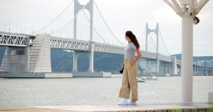 Thoughtful female tourist stands Busan's waterfront, looking down before raising head to cast farewell glance at suspension bridge over Seyeong Bay in background. Slow motion telephoto shot