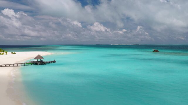 Aerial view of a Maldives resort lagoon. Camera glides over turquoise water, curved white sandbank, palms, wooden jetty with thatched pavilion, and a small floating hut.
