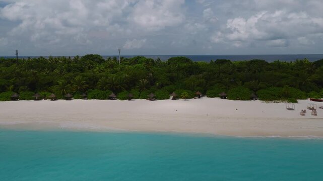 Aerial view shows a crescent white sand beach and turquoise lagoon with cabanas, umbrellas, a pavilion, lounge chairs, daybeds, and two telecom masts as waves lap