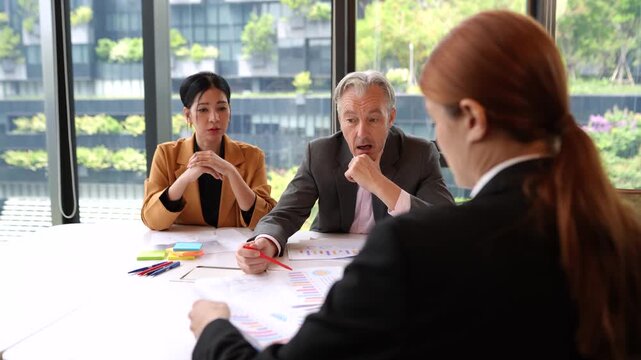 Over the shoulder shot of female employee while senior executive and secretary review financial report and business performance documents together during professional corporate office meeting.