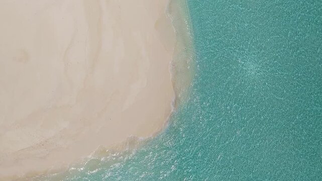 Aerial top down view shows a crescent sandbank and turquoise lagoon in the Maldives. Gentle waves lap, sand patterns shift, faint footprints mark bright daylight.