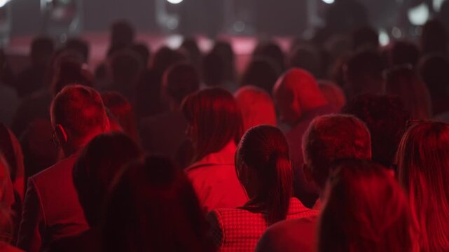 Audience seated in dim red light, silhouettes of people facing stage, focused atmosphere, conference or concert setting.