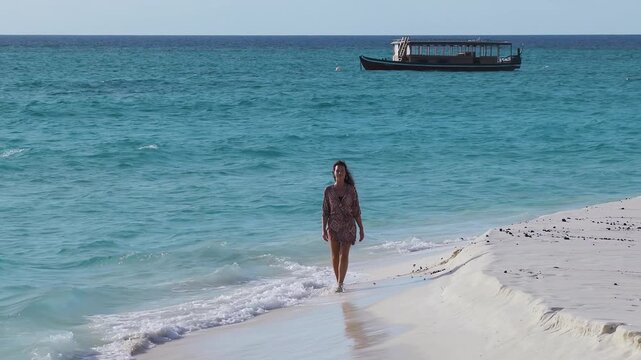 A woman walks barefoot on powder white sand as gentle waves lap. A Maldivian dhoni anchors offshore near thatched overwater villas in the Maldives at dusk.