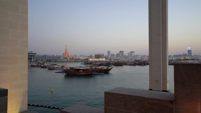 Framed shot from MIA Park overlooking Doha's historic harbor at dusk, featuring traditional dhow boats on calm waters with the iconic glowing Fanar spiral minaret and city skyline beyond.
