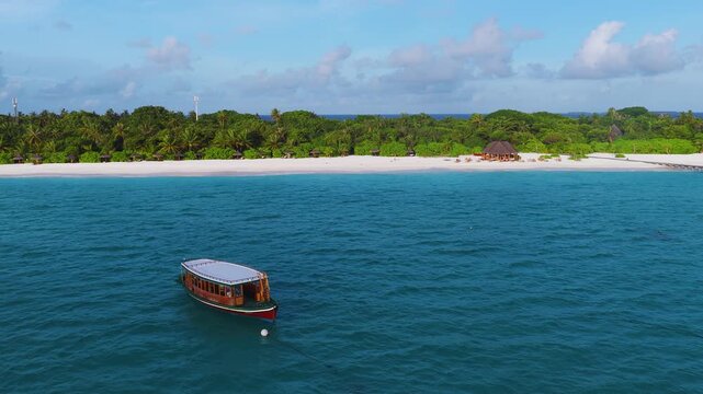 Aerial view shows a Maldivian resort island with white sand, palm cover, a thatched pavilion, a long wooden jetty, and a moored dhoni as the camera pans in warm sunlight.