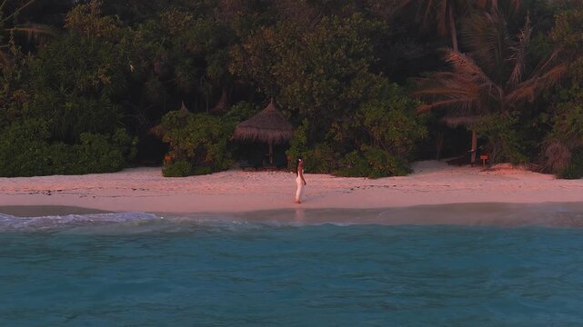 Aerial wide drone view shows Maldives white sand beach, palms, and palapas at sunset. A woman in a white dress walks as turquoise waves lap with steady motion.