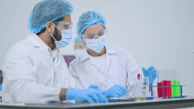Male and female researchers in protective masks, caps and gloves working at lab bench with test tubes of colored liquid, scientific teamwork.