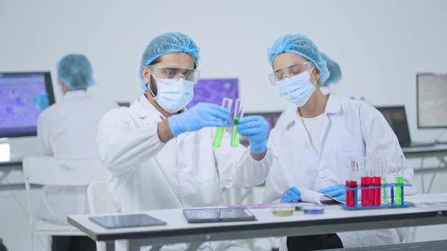 Male researcher in mask and goggles holding two test tubes with green chemical while female colleague watches, microscopy images on screens behind.