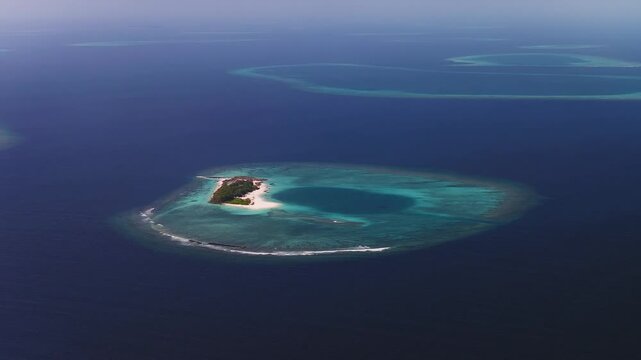 Aerial pan shows a ring shaped atoll in Maldives, a sandy palm islet in a turquoise lagoon, white surf on reef, dark ocean beyond, faint atolls on horizon in hazy daylight.