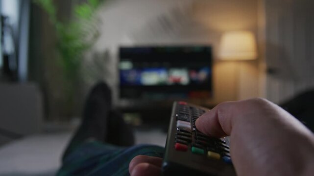 A man relaxes on the sofa, holding a remote control. He browses through channels on his smart TV, enjoying the comfort of his living room and the thrill of choosing entertainment