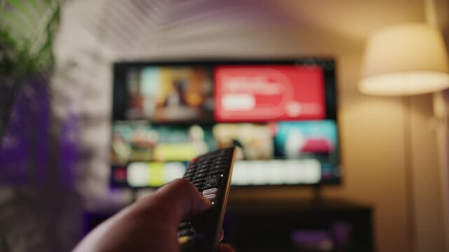 A man relaxes on his sofa, using a remote control to switch channels on a smart TV. The warm lighting and modern decor create a cozy atmosphere for entertainment