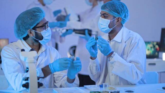 Male scientists in masks and protective caps holding small chip device at lab bench with microscope, technology research and development.