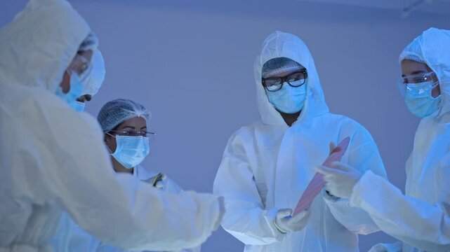 Four specialists wearing full body protective suits, masks and glasses talking during work, cleanroom production and laboratory safety concept.
