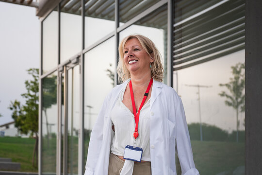 Senior female physician wearing a lab coat and id badge, smiling while walking outside a modern hospital building, representing healthcare professionalism and medical expertise