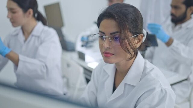 Young woman wearing eyeglasses looking at small specimen in gloved hand, research colleagues working in background, focused lab analysis.