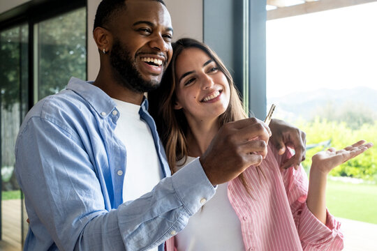 Happy couple holding house keys and smiling together in bright home by large window, celebrating new homeownership, love and future
