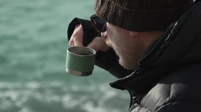 Man drinking coffee man in winter attire drinks a hot beverage from a thermos while standing near the ocean on a chilly day