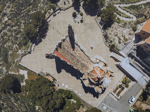 Aerial view of the Sanctuary of Santa Maria Magdalena at Mola Castle, featuring its unique modernist architecture, tiled roof, and surrounding paved plaza in Novelda, Comunidad Valenciana, Spain.