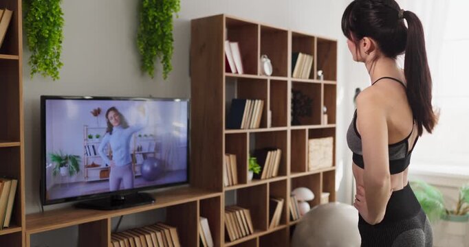 Woman workout stretching warmup fitness at home. Athletic female follows an online trainer on television, preparing to exercise in a cozy living room. Clear concept of modern home fitness.