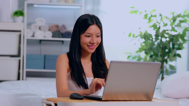 Young asian woman with laptop in bedroom at home smile while using computer online on bed, casual lifestyle workspace with plant shelf pillow natural light