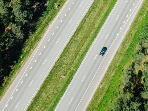 Aerial view of a single car driving on a multi-lane highway surrounded by lush green pine forests in Garkalne, Adazi Municipality, Latvia.