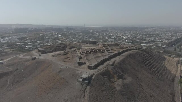 An aerial view beautifully showcases the ancient Erebuni Fortress, a historic Urartian fortified city situated on Arin Berd hill in Yerevan, Armenia, surrounded by the modern urban cityscape.

