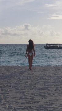A woman in a white bikini walks into turquoise surf in the Maldives. A Maldivian dhoni bobs offshore, atolls sit beyond, backlit in late afternoon light.