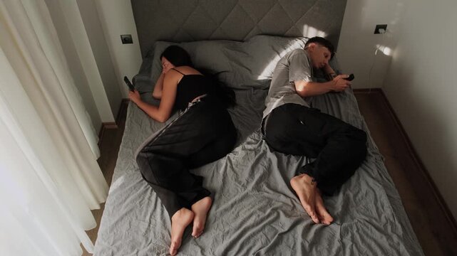 Phone addiction. Young couple ignoring each other while using smartphones in bed, relationship problems, emotional distance, digital distraction and lack of communication