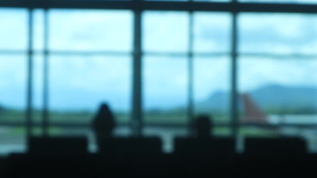 Blurry video of Silhouettes of children playing near chairs in a large airport waiting area before boarding, with a cloudy blue sky and views visible through panoramic glass windows. Makassar, Indones