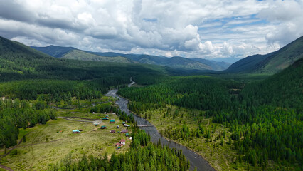 Scenic aerial panoramic view of a remote tourist base with small wooden cabins located in a green river valley surrounded by majestic Altai mountains. © OlTarakanov