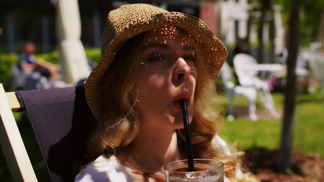 Woman sitting in a lounging chair a cafe in a green park outdoors and drinking a glass of iced coffee through a straw
