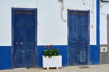 Fototapeta premium Two weathered dark blue wooden doors stand symmetrically on a white and blue painted facade with a central flower planter in Agaete, Gran Canaria.