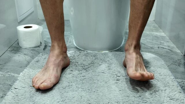 Close up view of a man's bare feet and lower legs while he sits on the toilet, restlessly moving and wiggling his toes out of boredom