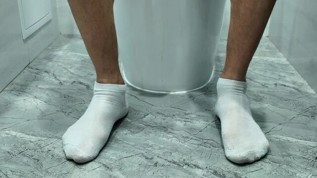A man's feet in white socks dance in boredom as he sits on a white toilet. The man reaches for toilet paper