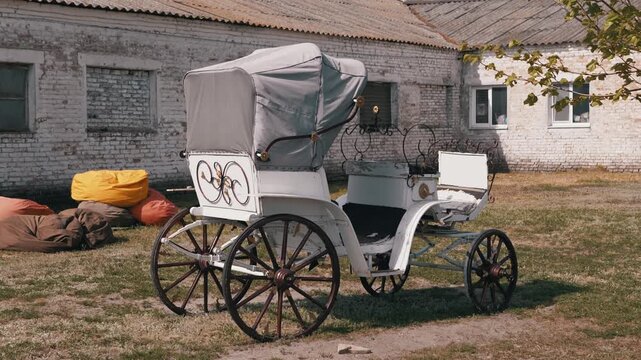 A white vintage horse carriage with a grey canopy sits on a grassy lawn before an old brick building. Concept of historical travel, romantic wedding transport, nostalgia, and rural heritage.